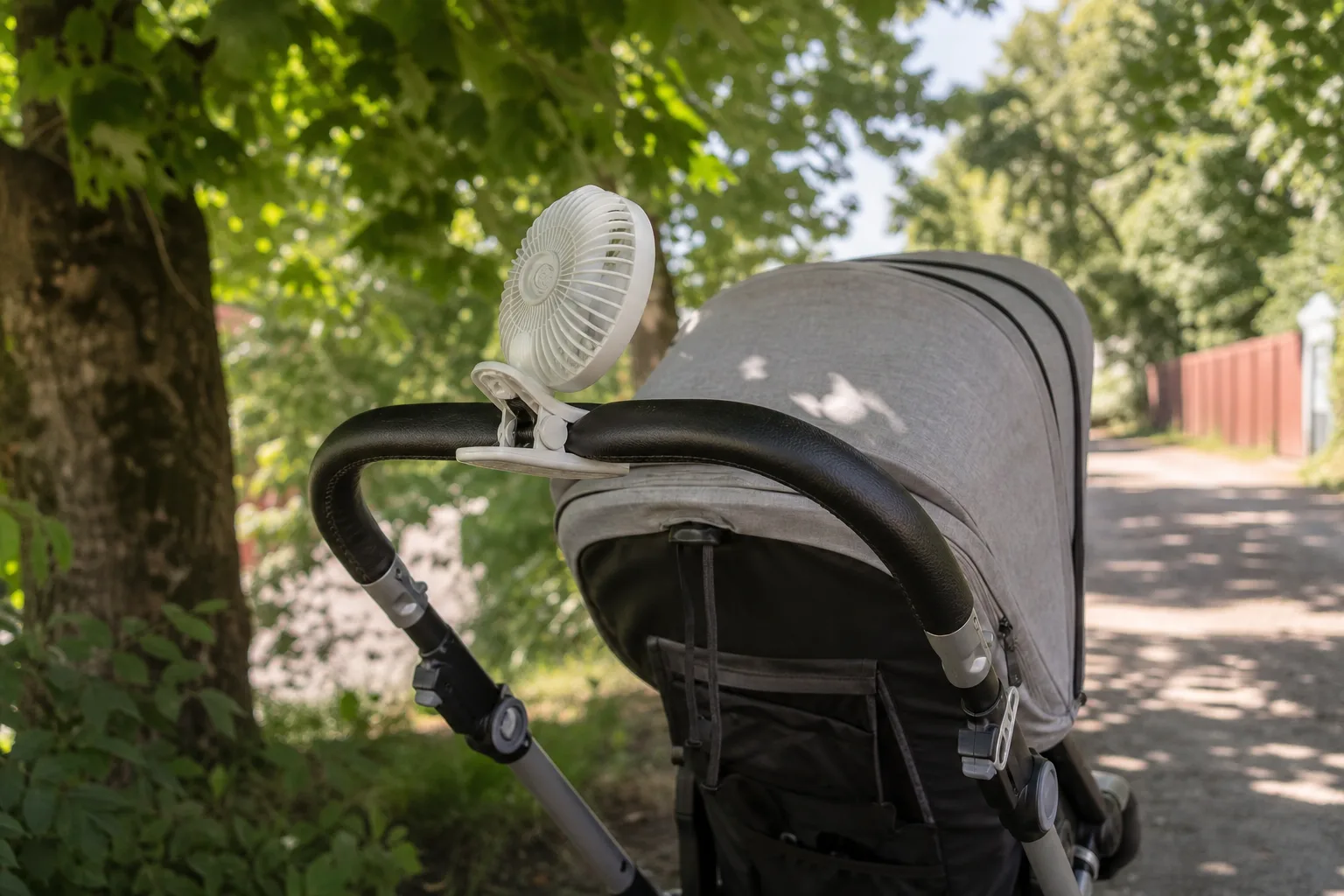 Vit klämfläkt fäst på styrhandtaget till en grå barnvagn som står i skuggan under ett träd